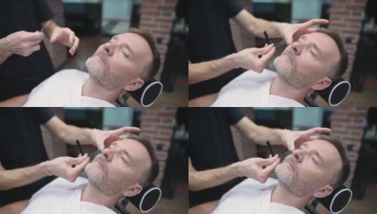 理发师大师在沙龙用电动剃须刀为英俊成熟的大胡子男子剃须。发型艺术家为男士理发店的人制作胡须造型。专业造型师服务。高清在线视频素材下载