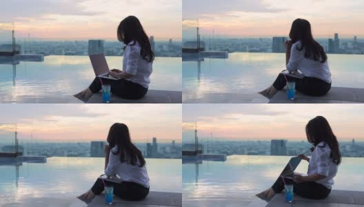 快乐的亚洲女人放松与笔记本电脑坐在边缘摇晃她的脚在游泳池看着远离梦想休息与城市的日落时间。高清在线视频素材下载