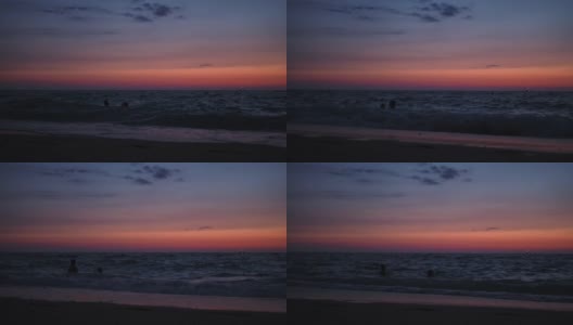 太阳落山后，人们在黄昏时到海里游泳。在远处，可以看到孩子们在波浪上跳跃。好玩的心情。红色的天空高清在线视频素材下载