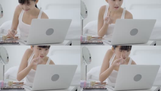 年轻的亚洲美女与学习化妆刷眼影笔记本电脑与教程课程在线，女性教学和解释眼影与化妆品在流媒体上化妆。高清在线视频素材下载