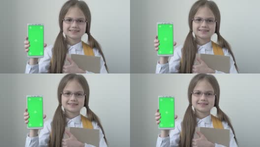 一名女学生，背书包，穿着白色的校衫，戴着眼镜拿着书，拿着绿色屏幕的手机。绿屏智能手机的色度键设置为广告。教育、技术和孩子高清在线视频素材下载
