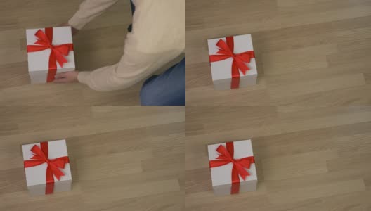 男子带来白色礼盒与红色丝带蝴蝶结，并把它放在地板上。木地板在室内。俯视图高角度。穿牛仔裤的男人拿着礼品盒走了。高清在线视频素材下载