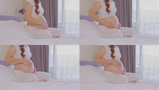 快乐怀孕的亚洲女人坐在床上抱着和抚摸着自己的大肚子，年轻女人的怀孕享受和未来的生活放松在家里，母亲和怀孕的概念，柔软的焦点高清在线视频素材下载