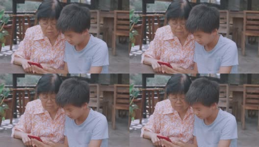 亚洲孙子教奶奶使用智能手机，帮助银发一代使用数字技术，社交距离家庭，多代概念高清在线视频素材下载