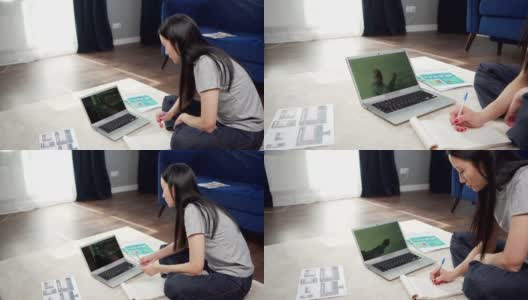 倾斜侧面的亚洲女大学生坐在家里的地板上，在线学习，观看视频会议与老师，并在笔记本上做笔记高清在线视频素材下载