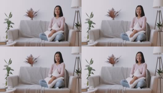 亚洲的美丽。年轻快乐的中国女士坐在家里的沙发上，抱着枕头对着镜头大笑高清在线视频素材下载