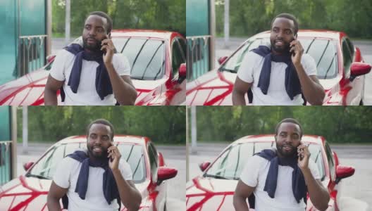 这张照片拍摄的是一位迷人的30岁非洲裔美国人，他面带微笑，留着整洁的胡子，穿着白色t恤，正在洗车时享受着在自己闪亮的红色汽车旁的电话交谈高清在线视频素材下载