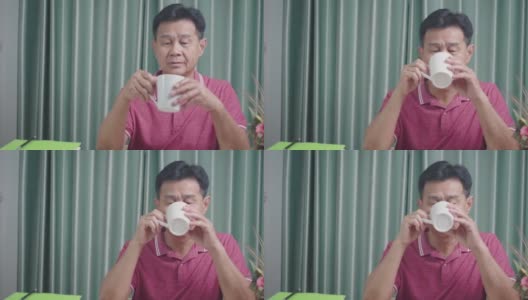 亚洲老男人从白色的杯子喝热饮，休息一天坐在家里的办公桌，退休生活，成熟的人类阶段，早上咖啡咖啡开始新的一天高清在线视频素材下载