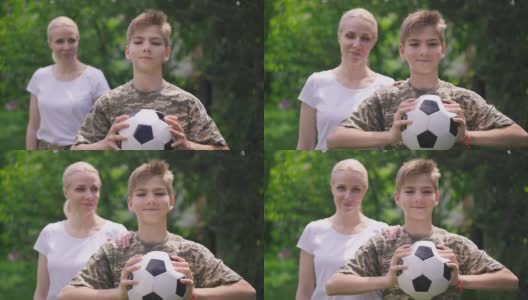 积极的十几岁的男孩摆姿势与足球，模糊的军事妇女把手放在肩膀上的慢动作的肖像。快乐自信的白人儿子和骄傲的母亲在公园摆姿势。高清在线视频素材下载