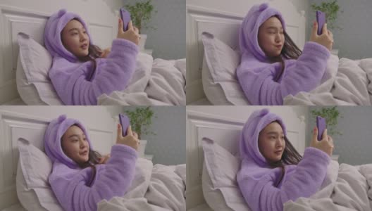 一名20多岁的亚洲女性躺在床上，一边看手机一边打视频电话。穿着紫色睡衣的漂亮女人在床上参加在线会议。高清在线视频素材下载