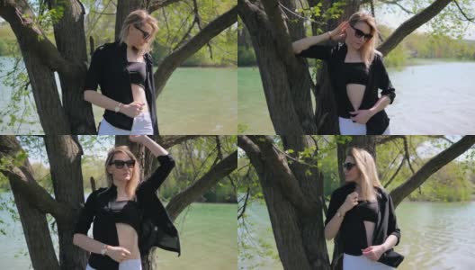 一位漂亮的年轻女子戴着太阳镜，穿着黑色的上衣和一件没有扣扣子的衬衫，站在树下，在湖边的大自然中拍照高清在线视频素材下载
