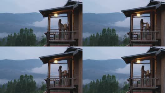 一个女人在木制房子的阳台上喝饮料，在雾天可以看到美丽的山景高清在线视频素材下载