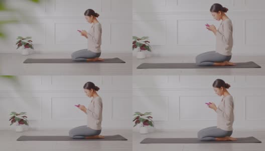 快乐的亚洲年轻女子坐在瑜伽垫放松与手机采取刹车后，与瑜伽练习在家里。瑜伽锻炼后休息的女人用手机发短信，面带微笑，心情愉悦高清在线视频素材下载