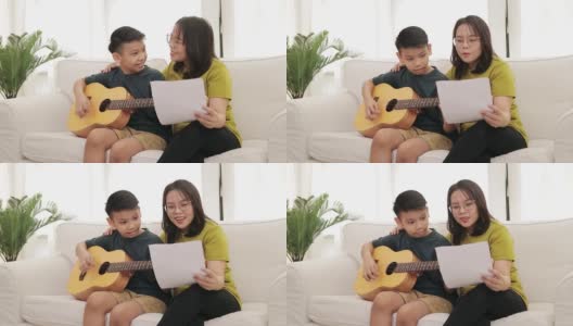 亚洲母亲和儿子坐在房子的白色沙发上儿子弹吉他，母亲唱歌高清在线视频素材下载