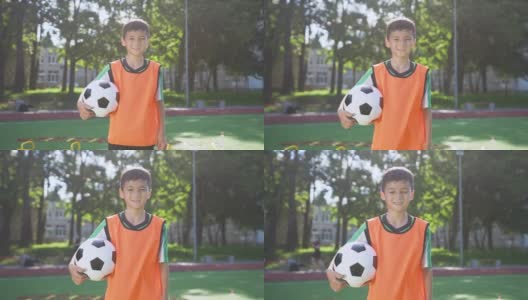 英俊疲惫的亚洲少年足球穿着站在球场与球之间的城市公园树木和严肃的脸看着相机高清在线视频素材下载