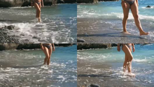 阳光明媚的日子里，年轻女孩的双腿沿着卵石海滩行走。温柔的海浪拍打着在海滩上赤脚行走的女性的脚。美丽的女人在比基尼享受夏天的旅行。近距离高清在线视频素材下载