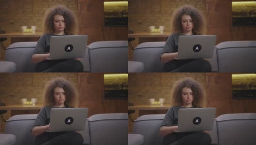 20多岁的卷发女人坐在家里的沙发上用笔记本电脑工作。年轻专注的女性在膝上的笔记本电脑上打字。高清在线视频素材下载