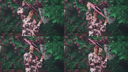 旧复古奥黛丽·赫本风格。粉红色的宽边帽子。自然化妆品概念，节日猫眼眼线化妆，可爱的脸性感的嘴唇。春天盛开的花园绿玫瑰丛中。时尚50 60年代高清在线视频素材下载