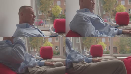 一个男人坐在椅子上，背靠背景窗户。他用手摸脸。高清在线视频素材下载