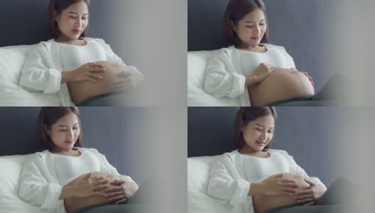 快乐的亚洲孕妇抚摸肚子，而躺在床上的卧室在家里。高清在线视频素材下载