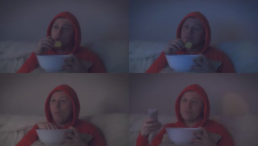 戴兜帽的男人在晚上看一部有趣的电影，从一个大碗里吃薯片，摄像机在移动高清在线视频素材下载