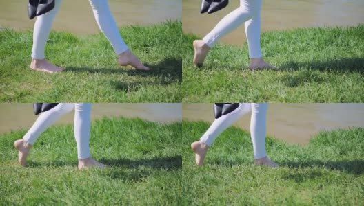 一个年轻女孩的脚赤脚走在绿色的草地在夏天一个阳光明媚的日子沿着湖。侧视图高清在线视频素材下载