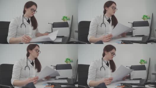 一名戴眼镜的年轻女医生通过视频通话技术与患者进行虚拟会面，她手里拿着患者的测试结果，并在诊所办公室的笔记本电脑上给出治疗建议高清在线视频素材下载