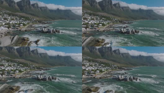 飞上海岸。汹涌的海浪冲击着海岸的岩石。高山峻岭下的住宅区。南非开普敦高清在线视频素材下载