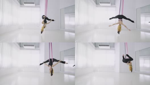 苗条运动年轻女子在黑色运动服展示瑜伽飞行在吊床上。一名年轻的亚洲女子在白色的体操室内进行反重力空中练习，手拿粉色丝带倒挂在空中。高清在线视频素材下载