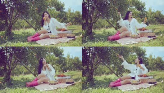 年轻的亚洲迷人的女性穿着休闲服装坐在放松野餐在橙新鲜的花园农场，女孩在野餐与三明治，水果和饮料。农场女孩吃午饭。高清在线视频素材下载