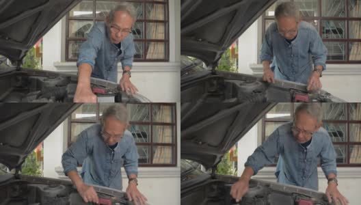 一位戴着眼镜、头发灰白的亚洲老人正在家门口检查一辆旧汽车的发动机部件状况。高清在线视频素材下载