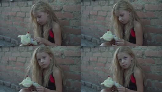一个来自贫困地区的悲伤小女孩靠着一堵砖墙坐在那里，手里拿着一个旧的填充玩具。高清在线视频素材下载