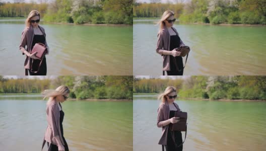 春天，一个年轻美丽的女人把一些东西放在她的手提包里，背景是城市公园里的一个湖高清在线视频素材下载