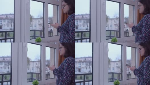 一个戴着眼镜，手里拿着手机的女孩站在窗口，用手机应用程序看外面，侧视图高清在线视频素材下载