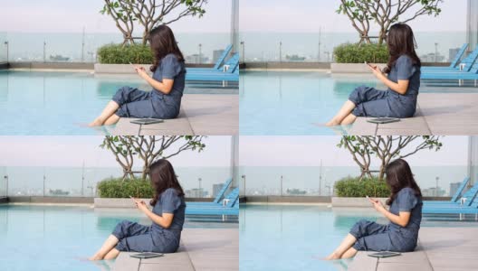 亚洲女性使用智能手机分享照片，度假时与朋友交流，坐在摩天大楼的游泳池边晃荡她的脚，放松时看着城市风景。高清在线视频素材下载