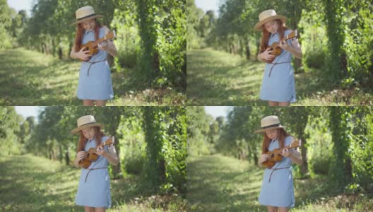 在春天的公园里，一个红发少女站在阳光下，穿着蓝色的衣服，戴着草帽，弹着四弦琴。有才华的自信的高加索青少年享受户外爱好的肖像。高清在线视频素材下载