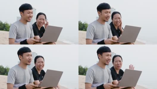一个留着胡子的女人和一个男人坐在沙滩上，手里拿着笔记本电脑。与朋友视频通话，并在一个明亮的日子在海边度假放松高清在线视频素材下载