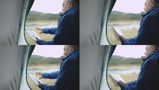 老人在帐篷里看书，在山前湖边露营——老人在大自然的概念下旅行高清在线视频素材下载