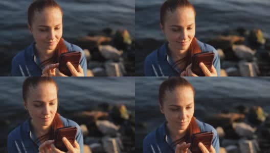 年轻漂亮的红发白人女子，手拿手机，坐在海边的一块大石头上，在社交网络上交流高清在线视频素材下载