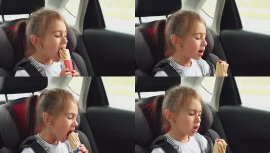 孩子在车里吃热狗。汽车后座上的女孩在旅途中被系上了安全带。高清在线视频素材下载
