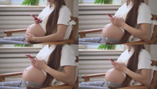 孕妇坐在家里的扶手椅上用手机和摸肚子高清在线视频素材下载