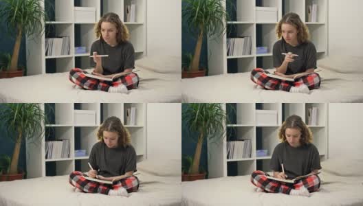 可爱的十几岁女孩坐在家里的床上做作业。一个年轻漂亮的女孩思考并在笔记本上写东西，在线学习，远程学习，自我教育。高清在线视频素材下载