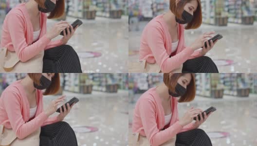 美丽的亚洲女性戴着防护口罩在商店使用智能手机，新常态概念，新冠肺炎后的人们生活方式高清在线视频素材下载