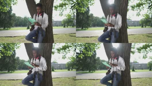 年轻专注的非洲裔美国学生倚在树干上用平板电脑上网。在阳光明媚的春天早晨，一名聪明的男子在户外检查作业。高清在线视频素材下载
