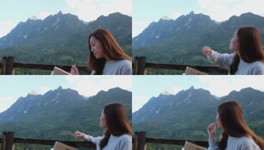 一位女游客坐在木制阳台上看书，吃零食，看着美丽的山景高清在线视频素材下载