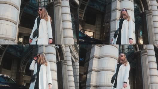 一个戴着眼镜，穿着白色外套的女孩走过城市的商业区。在豪华轿车和公寓的背景下高清在线视频素材下载