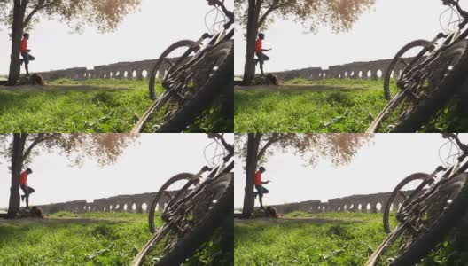 骑自行车的人站在一棵树下的岩石上，在古罗马的渡槽前看地图。年轻有吸引力的运动员与橙色运动装和背包parco degli acququedotti在罗马高清在线视频素材下载
