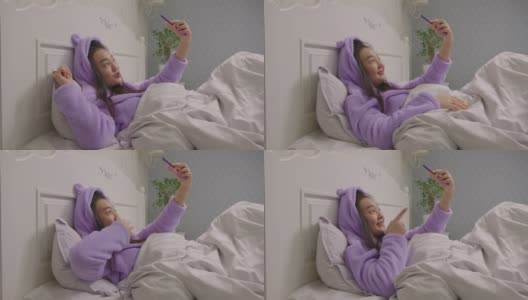 20多岁的亚洲女性躺在床上看着手机摄像头说话。穿着紫色睡衣的漂亮女博主在床上视频通话。高清在线视频素材下载