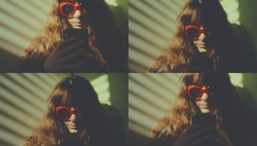 透过百叶窗的光线，映入眼帘的是一个戴着红色太阳镜的穿孔女孩，她正在用智能手机冲浪。慢动作拍摄高清在线视频素材下载