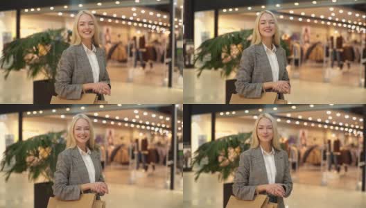 迷人的时尚年轻女子的肖像，穿着时尚的衣服站在轻购物中心，拿着纸袋看着相机。高清在线视频素材下载